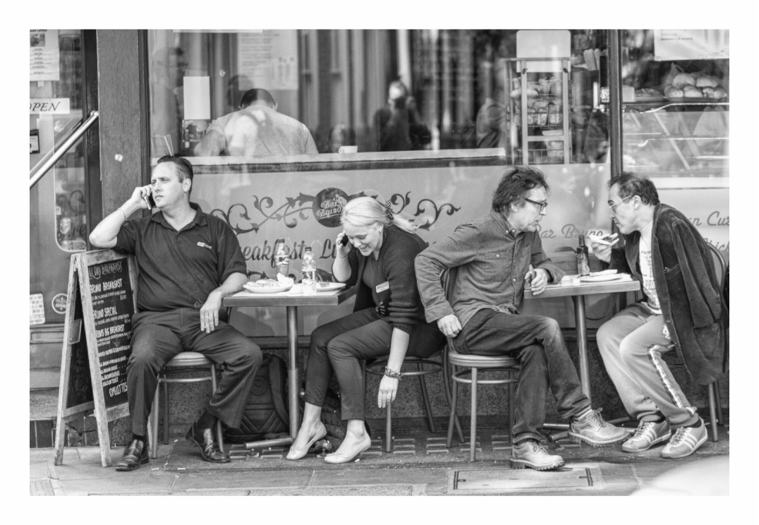 The Art Of Conversation print, 120 × 80 cm with white border — four people seated outside a Soho café, two on their phones, black and white London street photography by Tom Parsons