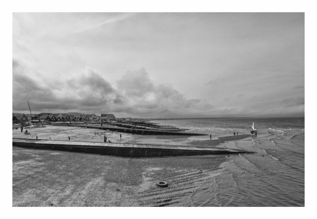 Whitstable print, 120 × 80 cm with white border — view from the harbour across the beach and jetties toward the sea, black and white Kent seafront photography by Tom Parsons
