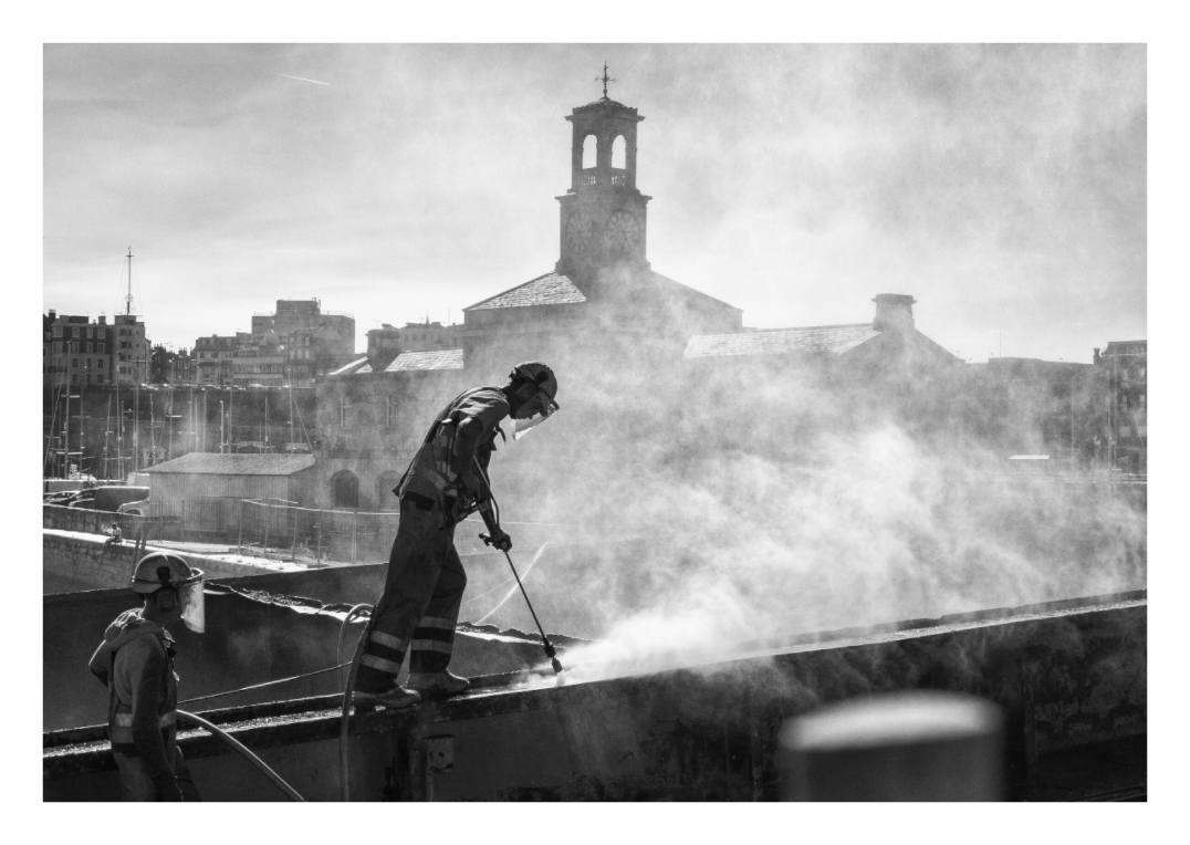 Boat worker print, 130.9 × 90 cm with white border — worker jetwashing a barge hull in steam and spray, harbour church behind, Ramsgate, Kent, black and white documentary photography by Tom Parsons
