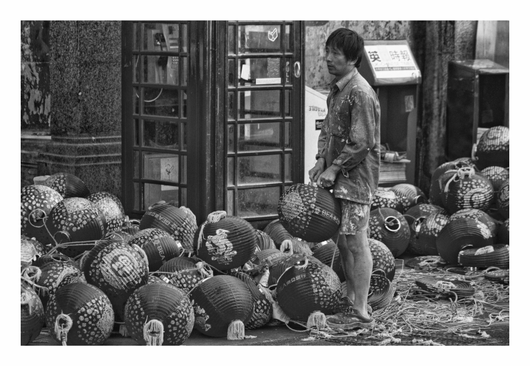 China Town print, 120 × 80 cm with white border — man dwarfed by a mountain of paper lanterns before Chinese New Year, black and white London street photography by Tom Parsons