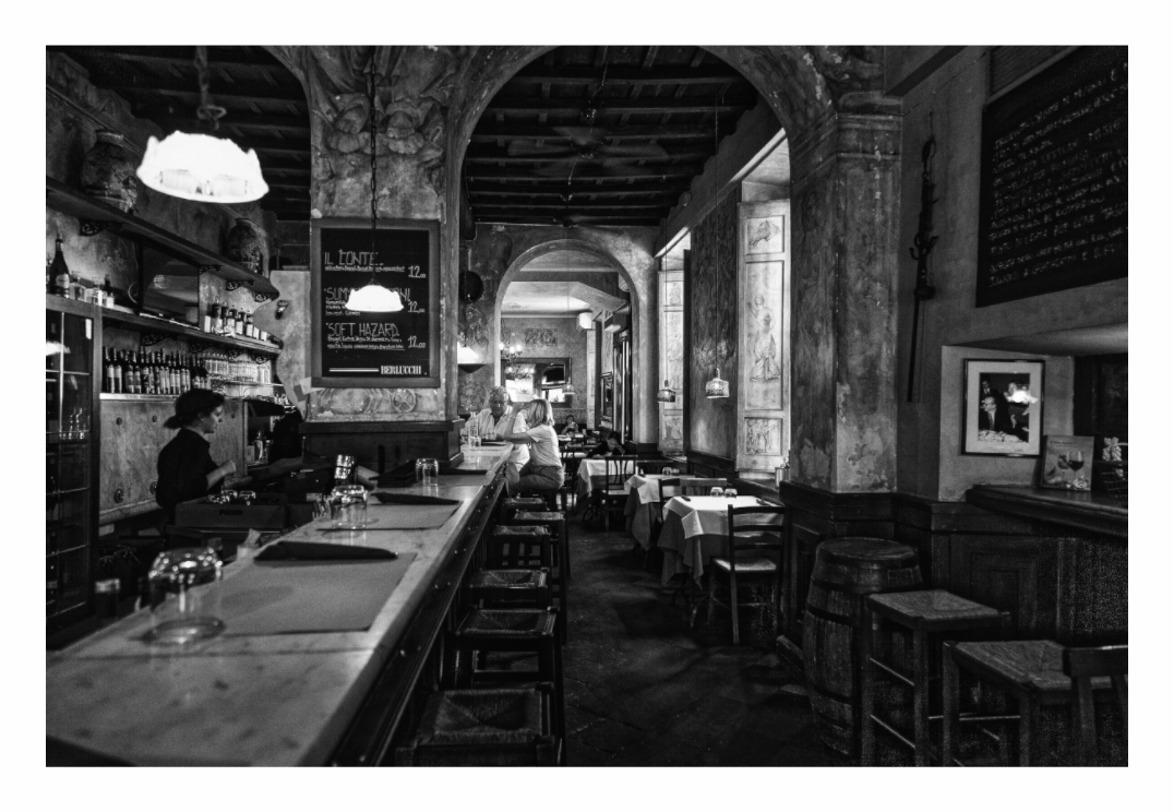 Bar in Rome print, 120 × 80 cm with white border — worn stone arches, a bartender at work, a woman seated alone at the counter, black and white Italian bar interior photography by Tom Parsons