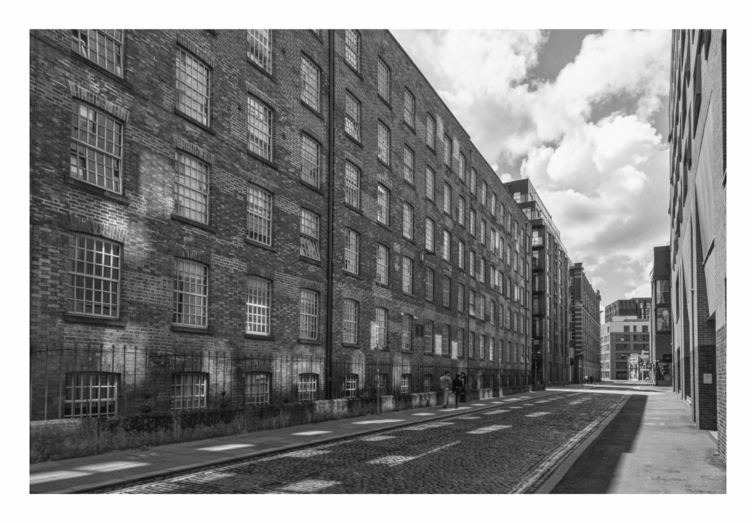 Manchester Street print, 120 × 80 cm with white border — long Victorian brick warehouse facade along a Manchester street, distant figures and modern buildings beyond, black and white street photography by Tom Parsons
