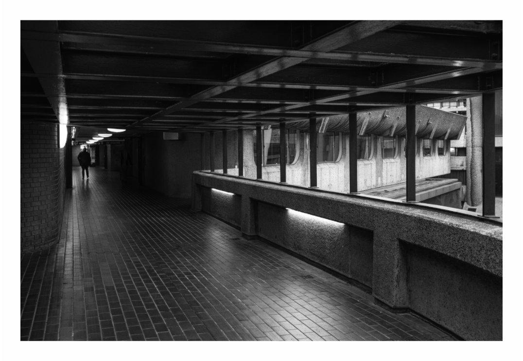 Barbican print, 120 × 80 cm with white border — lone figure walking through a covered walkway at the Barbican Estate, London, black and white architecture photography by Tom Parsons