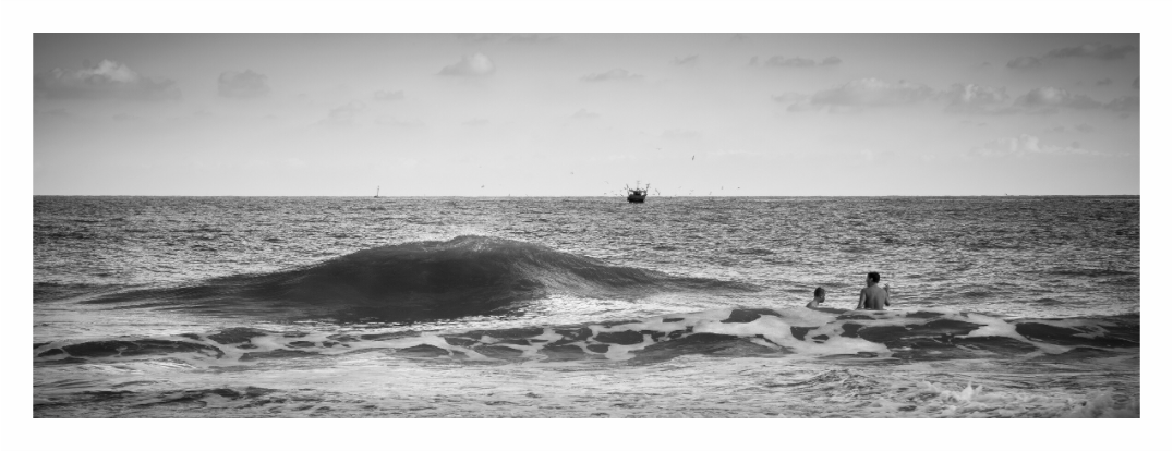 The Sea print, 172.1 × 60 cm with white border — two figures sitting in the surf at Ramsgate beach, Kent, with a boat passing behind, black and white panoramic seascape photography by Tom Parsons