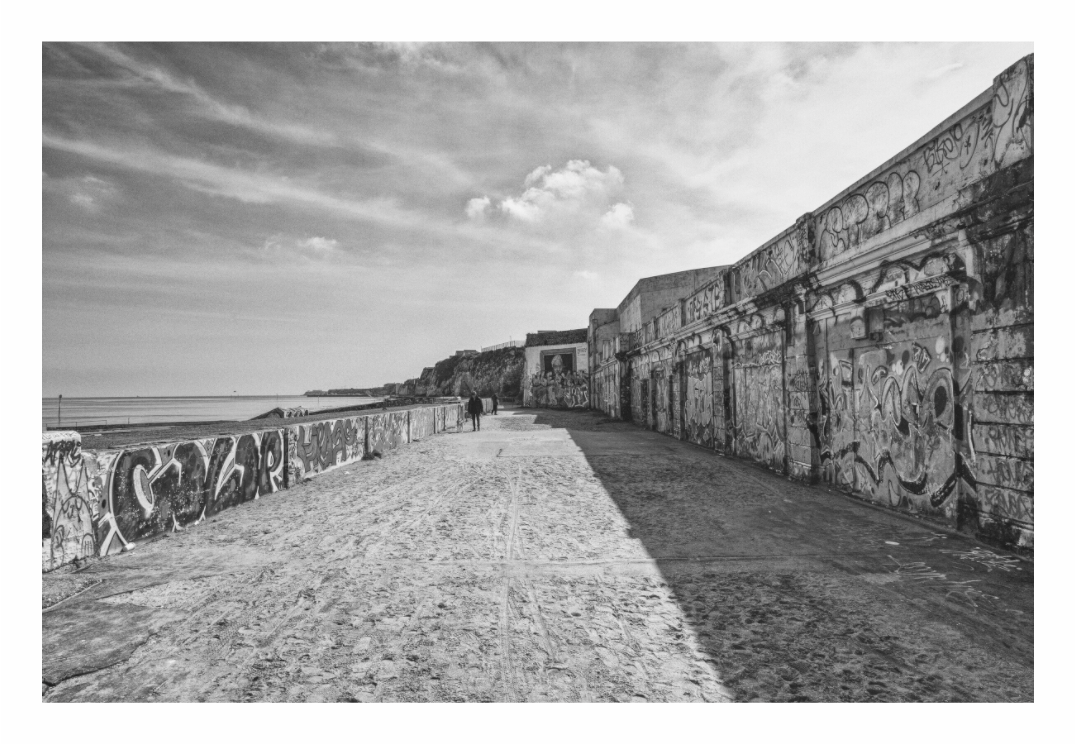Margate print, 120 × 80 cm with white border — graffiti-covered sea wall along the lower promenade, two figures walking in the distance, black and white Margate seafront photography by Tom Parsons