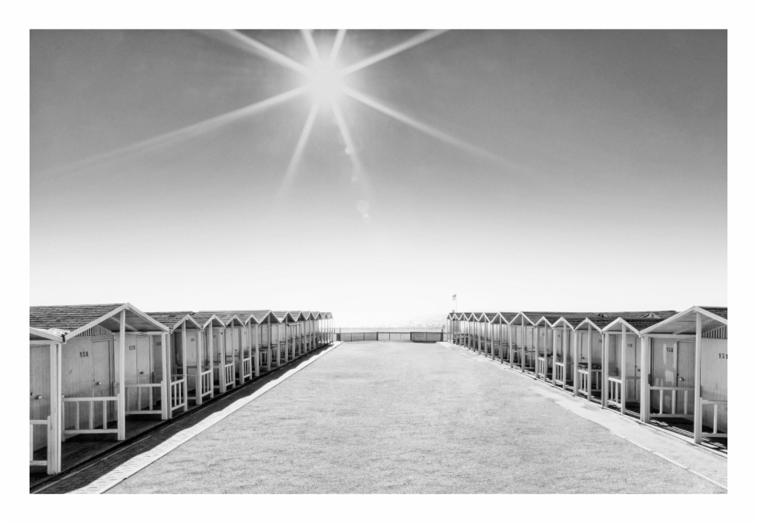 Beach huts print, 120 × 80 cm with white border — symmetrical rows of beach huts with a bright sunburst overhead, Ostia, Lazio, Italy, black and white landscape photography by Tom Parsons