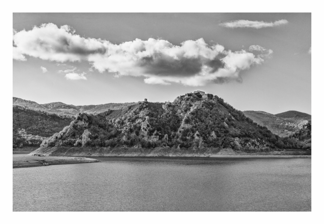 Castel di Tora and lake Turano print, 120 × 80 cm with white border — Castel di Tora perched on a rocky hill above Lake Turano beneath a dramatic cloudscape, Lazio, Italy, black and white landscape photography by Tom Parsons