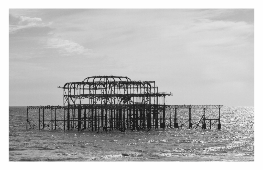 Brighton Pier print, 144.4 × 90 cm with white border — ruined skeletal ironwork of Brighton's West Pier standing in the sea, black and white architecture photography by Tom Parsons