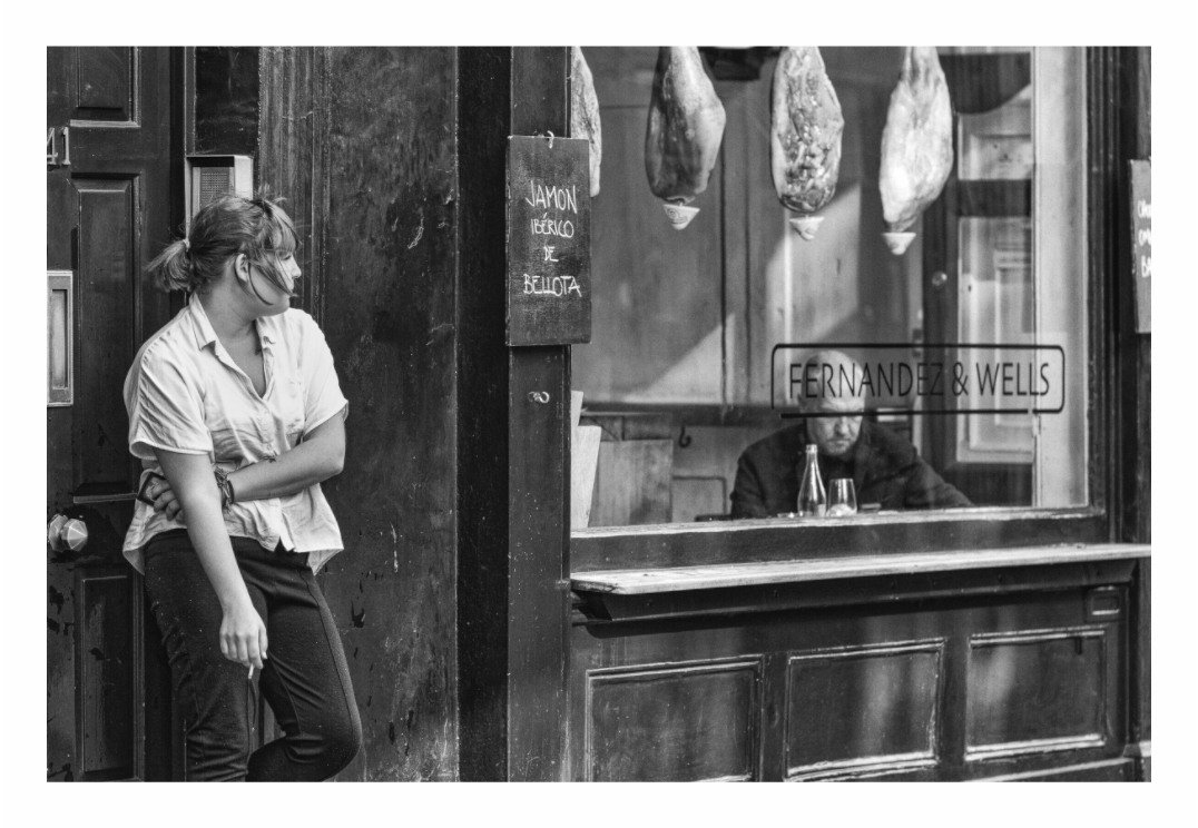 The Window print, 120 × 80 cm with white border — woman leaning outside a Soho deli, man seated behind glass among hanging hams, black and white London street photography by Tom Parsons