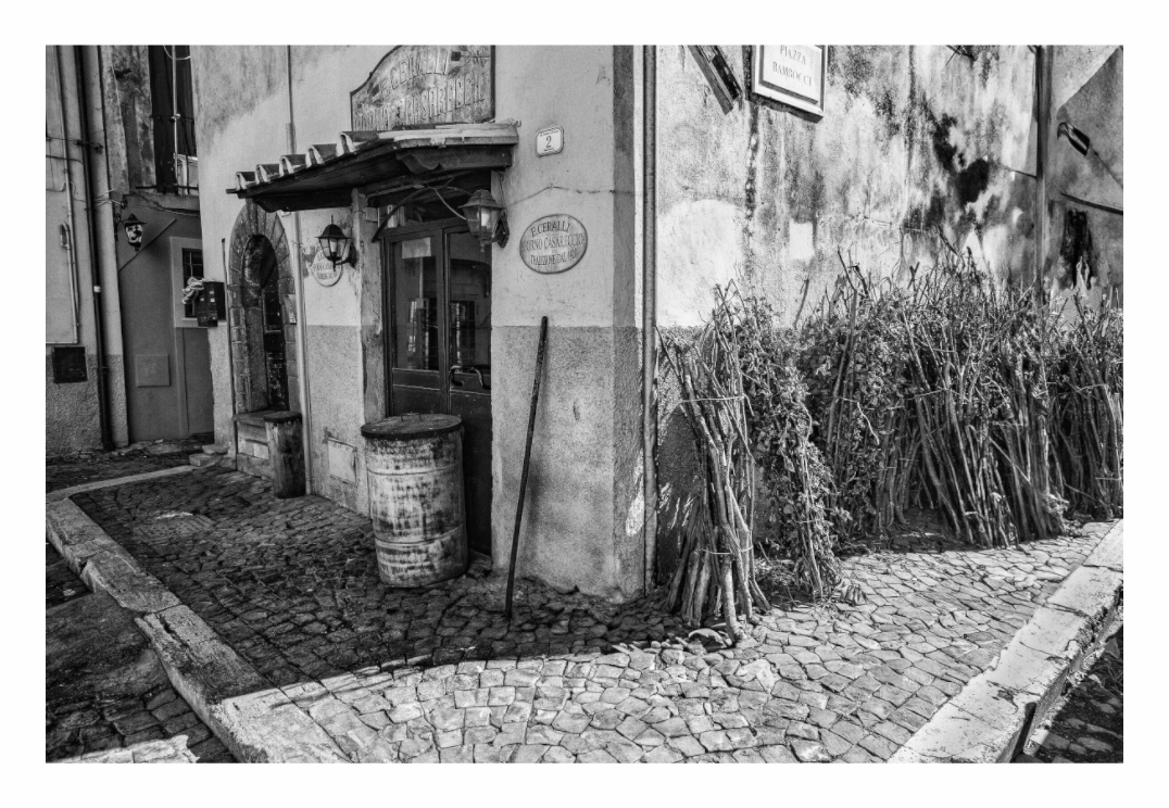 Street Corner print, 120 × 80 cm with white border — cobblestone street corner with a rustic doorway and bundled reeds, Italy, black and white street photography by Tom Parsons