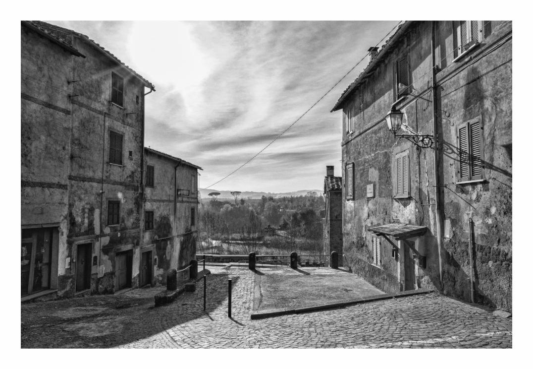 Italian Street print, 120 × 80 cm with white border — a cobbled street corner in a hilltop Italian village, countryside beyond, black and white street photography by Tom Parsons