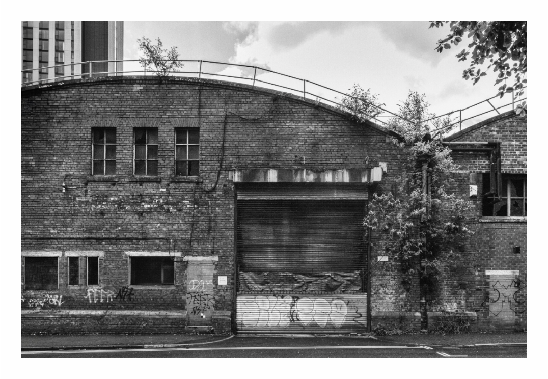 Abandoned Warehouse print, 120 × 80 cm with white border — shuttered, graffiti-covered loading bay under a curved ivy-grown roof, tower block behind, Manchester, black and white architecture photography by Tom Parsons